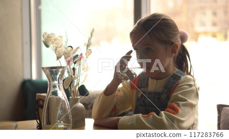 A girl drinks water in a cozy cafe, sitting at a table with a panoramic window in the background. A girl drinks water in a cozy cafe, sitting at a table with a panoramic window in the background. 117283766