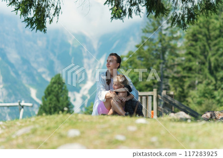 Attractive young mother and her cute daughter are sitting under a tree in the shade on mountain peak Attractive young mother and her cute daughter are sitting under a tree in the shade on mountain peak 117283925