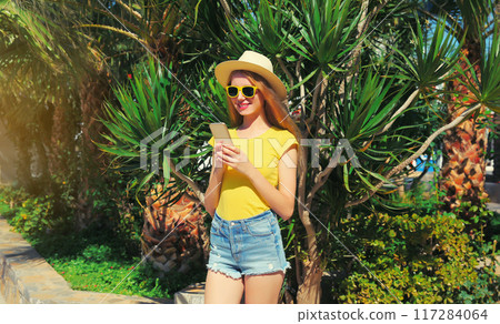 Portrait of young woman with smartphone in summer park on palm tree background Portrait of young woman with smartphone in summer park on palm tree background 117284064