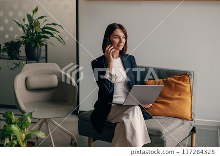A businesswoman is talking on the phone with a laptop in a modern office surrounded by plants 117284328