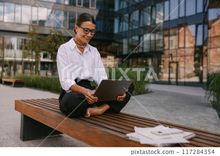 A professional woman is working outdoors while using her laptop in a relaxed setting A professional woman is working outdoors while using her laptop in a relaxed setting 117284354