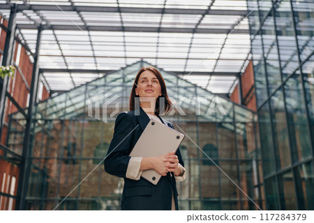 A confident businesswoman is standing in modern glass architecture, holding documents 117284379