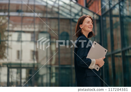 A successful businesswoman confidently holds a laptop in front of a contemporary office building A successful businesswoman confidently holds a laptop in front of a contemporary office building 117284381