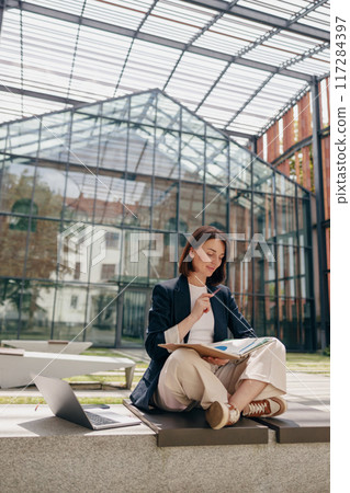 A woman is working outdoors with a laptop and notebook, relaxing in a courtyard 117284397