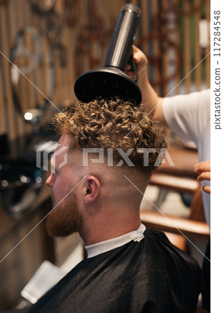 Professional woman-hairdresser is drying the hair of a curly red-haired man with hairdryer. 117284548