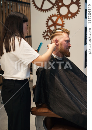 Red-haired bearded man sitting in barbershop and female barber making hairdo to guest 117284549