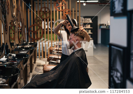 Professional woman-hairdresser is drying the hair of a curly red-haired man with hairdryer. 117284553