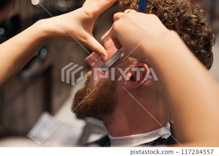 Female barber shaves a man's beard with a straight razor. Man during beard shaving in barber shop. 117284557