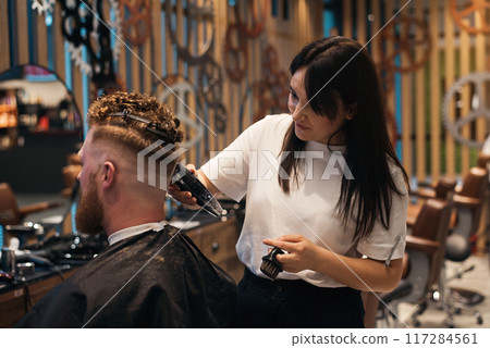 Red-haired bearded man sitting in barbershop and female barber making hairdo to guest 117284561