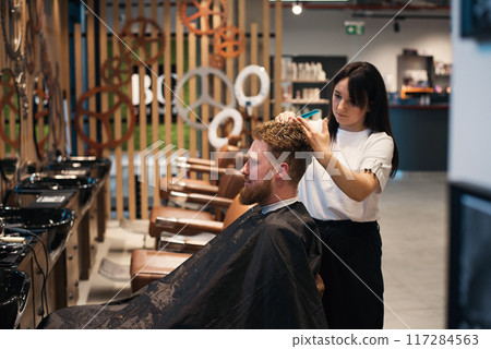 Red-haired bearded man sitting in barbershop and female barber making hairdo to guest 117284563