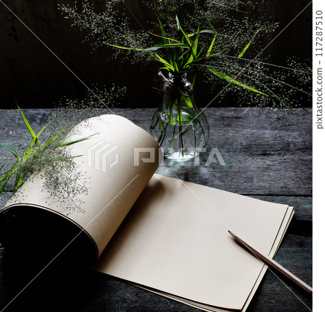 A still life scene, open sketchbook with a warm paper page, a pencil, and glass bottles holding flowers grasses, abstract shadow make creative expression, simple decor workspace 117287510