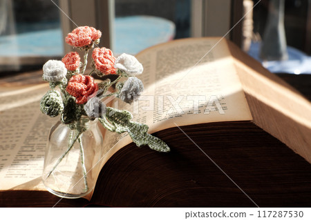 An open book rests beneath a small bouquet of delicate white flowers in a glass vase on rustic wooden surface, peaceful reading nook near window in vintage aesthetic An open book rests beneath a small bouquet of delicate white flowers in a glass vase on rustic wooden surface, peaceful reading nook near window in vintage aesthetic 117287530