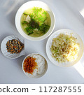 Vietnamese meal with steamed rice, tofu and vegetable soup, stir-fried green beans, bamboo shoots, and chili sauce on a white table, illuminated by natural light from a window. 117287555