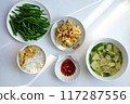 Vietnamese meal with steamed rice, tofu and vegetable soup, stir-fried green beans, bamboo shoots, and chili sauce on a white table, illuminated by natural light from a window. 117287556