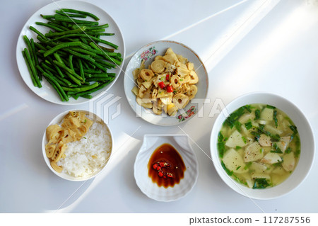 Vietnamese meal with steamed rice, tofu and vegetable soup, stir-fried green beans, bamboo shoots, and chili sauce on a white table, illuminated by natural light from a window. Vietnamese meal with steamed rice, tofu and vegetable soup, stir-fried green beans, bamboo shoots, and chili sauce on a white table, illuminated by natural light from a window. 117287556