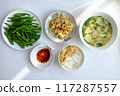 Vietnamese meal with steamed rice, tofu and vegetable soup, stir-fried green beans, bamboo shoots, and chili sauce on a white table, illuminated by natural light from a window. 117287557