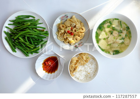 Vietnamese meal with steamed rice, tofu and vegetable soup, stir-fried green beans, bamboo shoots, and chili sauce on a white table, illuminated by natural light from a window. 117287557