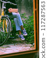 A young woman in jeans and sneakers sits on a yellow bicycle, holding a brick to troll moments, framed by a window with lush greenery behind, make humor and funny atmosphere 117287563