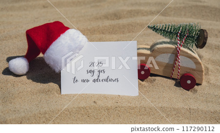 2025 SAY YES TO NEW ADVENTURES text on paper greeting card on background sandy beach sun coast. Christmas balls Santa hat New Year New Me Resolutions decoration. Summer vacation decor. Holiday concept 117290112