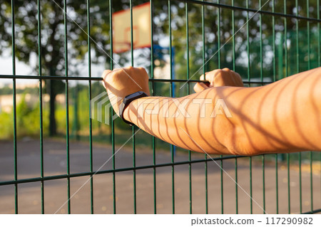 Close-up of a person s hand gripping a green metal fence, with a blurred background of trees and a basketball court. Represents strength, security, and boundaries. Close-up of a person s hand gripping a green metal fence, with a blurred background of trees and a basketball court. Represents strength, security, and boundaries. 117290982