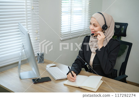 A woman wearing a hijab headset is talking on the phone while sitting at a desk A woman wearing a hijab headset is talking on the phone while sitting at a desk 117292274