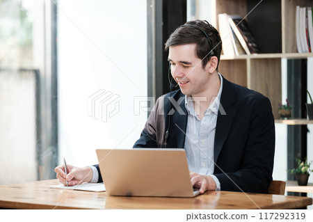 A man is sitting at a desk with a laptop and a pen 117292319