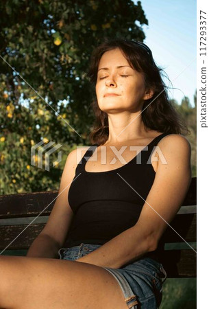 Young woman in casual clothes sitting on the bench in park in sunlight. Girl with closed eyes resting outdoor. Tranquility in big city lifestyle. Life balance concept. Meditation moment. Young woman in casual clothes sitting on the bench in park in sunlight. Girl with closed eyes resting outdoor. Tranquility in big city lifestyle. Life balance concept. Meditation moment. 117293377