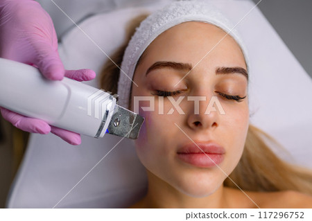 Facial cleansing with ultrasound scrubber. Woman receiving ultrasound facial peeling and cleansing. Cosmetology and face skin care. Facial treatment Facial cleansing with ultrasound scrubber. Woman receiving ultrasound facial peeling and cleansing. Cosmetology and face skin care. Facial treatment 117296752