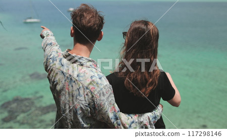 Young man is pointing to something on the horizon while hugging his girlfriend, both admiring the turquoise water and the sailboats in the distance 117298146