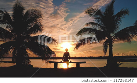Watching the sunset by the sea, a woman sits on a bench between palm trees, surrounded by nature's beauty in a tranquil tropical paradise 117298147