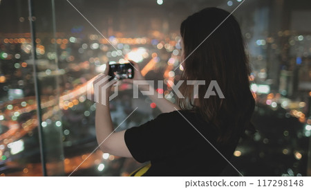 Woman tourist making a panorama of night city. Colorful cityscape center downtown, night lights view from top floor of Baiyoke Tower Sky, Bangkok, Thailand. Travel, tourism, slow motion 4K. Back view 117298148
