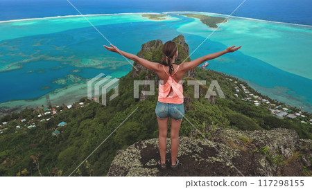 Young tourist stands on a mountain top. Arms outstretched. Enjoying the view of a tropical island with a turquoise lagoon below Young tourist stands on a mountain top. Arms outstretched. Enjoying the view of a tropical island with a turquoise lagoon below 117298155