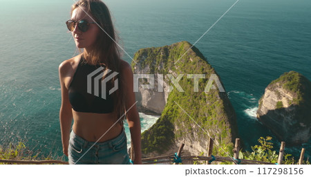 Young woman is taking in the view of the beautiful blue water and green cliffs of kelingking beach from a scenic viewpoint Young woman is taking in the view of the beautiful blue water and green cliffs of kelingking beach from a scenic viewpoint 117298156