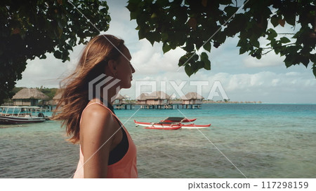 Tourist girl enjoy Bora Bora water villa bungalows sea view. Caucasian female standing on beach under the tree back view. Outdoor lifestyle travel on summer holiday vacation, French Polynesia 117298159