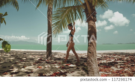 Woman in bikini walking sand tropical beach in Thailand, palm trees and clear blue sky. White sand beach and ocean in background. Girl relax outdoor lifestyle travel on summer holiday vacation. 117298174