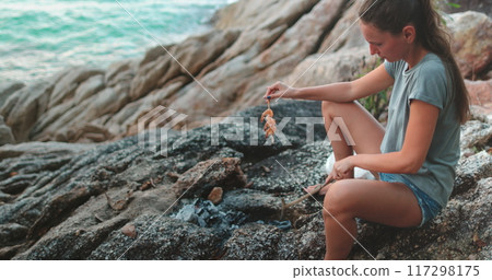 Grilling shrimp skewers on a beach campfire at sunset, a woman enjoys a tranquil evening by the sea 117298175