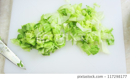Fresh iceberg salad cutting close up on white kitchen board. Chef hands in blue gloves mince green cabbage with big iron knife. Cooking, preparing ingredients in professional restaurant. Top down view Fresh iceberg salad cutting close up on white kitchen board. Chef hands in blue gloves mince green cabbage with big iron knife. Cooking, preparing ingredients in professional restaurant. Top down view 117298177