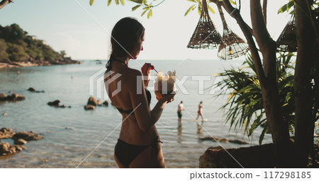 A woman stands on a beach in Thailand, holding a coconut cocktail drink in her hand. Sunset sea coastline palm trees. Female tourist on summer holiday vacation journey. Lifestyle relax concept A woman stands on a beach in Thailand, holding a coconut cocktail drink in her hand. Sunset sea coastline palm trees. Female tourist on summer holiday vacation journey. Lifestyle relax concept 117298185