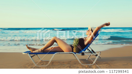 Summer vacation, happy relaxing young woman lying on deckchair with pineapple on the beach on sea 117299341