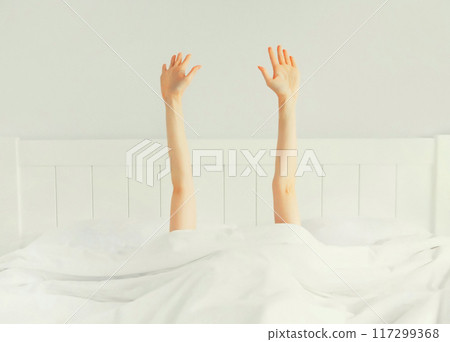 Lazy woman waking up after sleeping lying in bed showing gesture her hands up from under the blanket 117299368