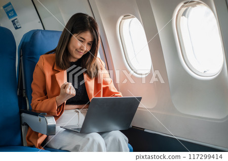 A young businesswoman working on her laptop while traveling on an airplane, showing focus and determination. A young businesswoman working on her laptop while traveling on an airplane, showing focus and determination. 117299414
