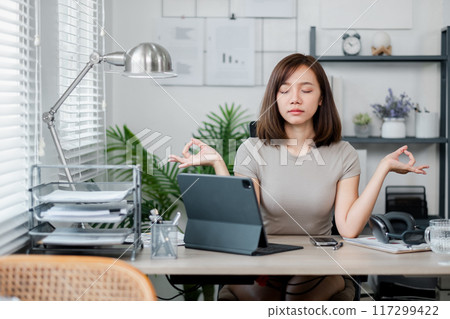 A young woman sits at her home office desk, practicing mindfulness meditation with a digital tablet in a modern workspace. 117299422