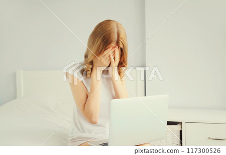 Portrait of sad upset crying young woman working with laptop on the bed in white room at home 117300526