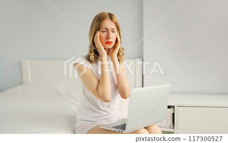 Portrait of tired young woman with headache worked with laptop on the bed in white room at home Portrait of tired young woman with headache worked with laptop on the bed in white room at home 117300527