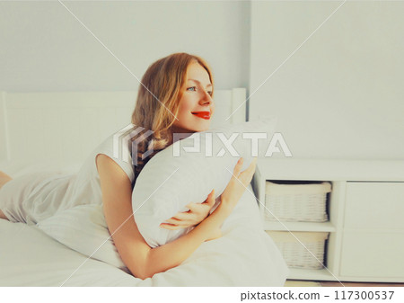 Healthy sleep, happy relaxed young woman hugs her pillow during lying on soft comfortable bed 117300537