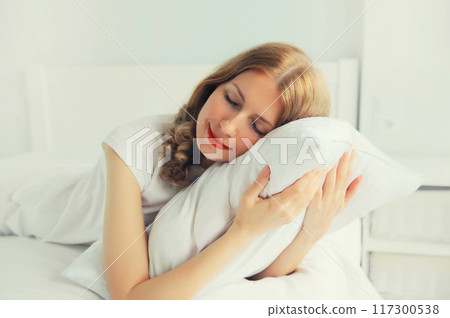 Healthy sleep, happy relaxed young woman hugs her pillow during lying on white comfortable bed 117300538