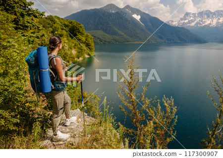 A hiker stands peacefully by a serene lake surrounded by majestic, towering mountains 117300907