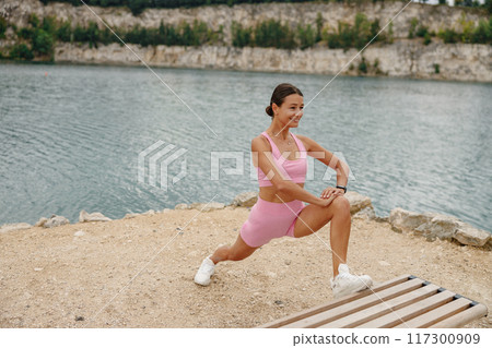 A woman is exercising outdoors by the clear water while wearing a stylish pink workout outfit A woman is exercising outdoors by the clear water while wearing a stylish pink workout outfit 117300909