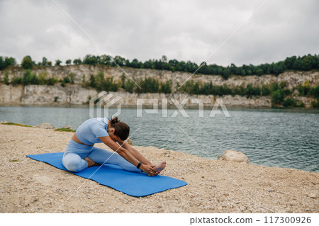 A woman is seen practicing yoga surrounded by the serene and picturesque environment of a lake A woman is seen practicing yoga surrounded by the serene and picturesque environment of a lake 117300926