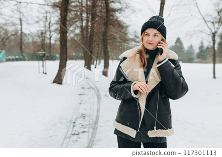 Beautiful girl on the winter snowy street with the phone. A blonde woman talking on a mobile phone outdoors. Technology, communication and people concept. 30s female speak phone on nature background, 117301113